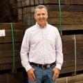 Professional portrait of a man standing in front of a stack of hardwood timber. He is smiling and has his thumbs in his pockets. He is wearing a light blue striped button-up and dark blue jeans with
