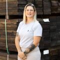professional portrait of a woman standing in front of a stack of timber products. She is wearing a grey shirt and beige formal pants