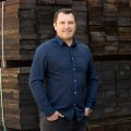 Professional man standing in front of a stack of hardwood timber with his hands in his pockets. he is wearing a dark blue formal button-up
