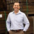 Professional man standing in front of a stack of hardwood timber smiling broadly. he is wearing a light blue formal button-up